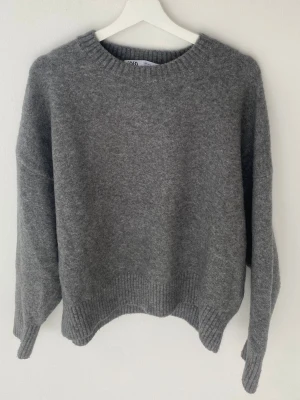 Grå stickad tröja från Zara - Mysig grå stickad tröja från Zara med rund halsringning och ribbade muddar vid ärmslut och nederkant. Tröjan har en avslappnad passform och är perfekt för kyliga dagar. Materialet känns mjukt och skönt mot huden.