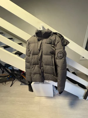 Canada Goose Mörkgrå pufferjacka med luva - Snygg mörkgrå pufferjacka med stor luva och quiltad design. Jackan har en patch på ärmen och dragkedja framtill med tryckknappar. Perfekt för kalla vinterdagar och ger en riktigt trendig look.