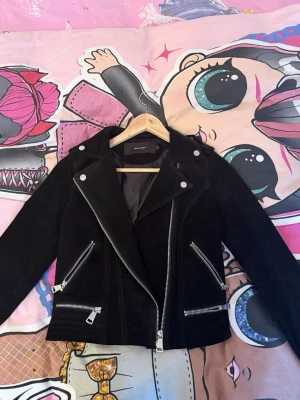 Svart skinnjacka från Vero Moda - Snygg svart skinnjacka från Vero Moda med silvriga dragkedjor och knappar. Klassisk bikerstil med bred krage och tre framfickor med dragkedja. Kortare modell som ger en cool och trendig look.