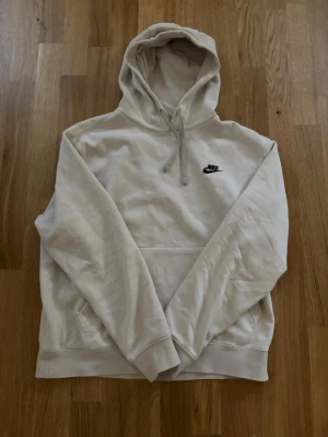 Beige hoodie från Nike med logga - Säljer en beige hoodie från Nike med svart broderad logga på bröstet. Hoodien har klassisk huva med dragsko, magficka och ribbade muddar vid ärmar och nederkant. Tillverkad i mjukt bomullsmaterial för skön känsla.