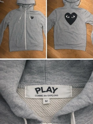 Grå hoodie från Comme des Garçons Play - Grå hoodie från Comme des Garçons Play med dragkedja, vit dragsko och ikoniskt svart hjärta med ögon på bröstet och ryggen. Tillverkad i mjuk bomullsblandning och har en klassisk huva. Perfekt för en clean och stilren look. Säljer pga att den inte passar längre (är 190cm lång) hör av er vid frågor☺️