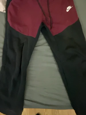 Nike tvåfärgade mjukisbyxor - Säljer ett par mjukisbyxor från Nike med vinröd överdel och svarta ben. Byxorna har dragsko i midjan och Nike-logga på vänster sida. Tillverkade i mjukt material som passar perfekt för chill eller träning. Snygg kontrastdesign och bekväm passform.