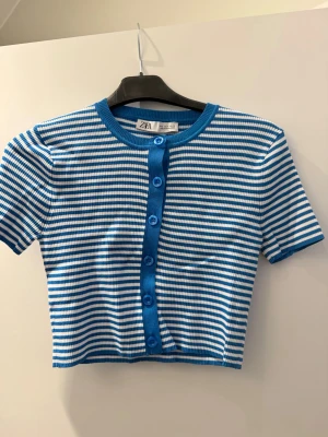 Randig blåvit croppad topp från Zara - Säljer en croppad topp från Zara med blå och vita ränder. Toppen har korta ärmar, rund halsringning och knappar framtill i matchande blå färg. Materialet är ribbat och stretchigt, vilket ger en bekväm passform. Perfekt för dig som gillar en fräsch och trendig stil.