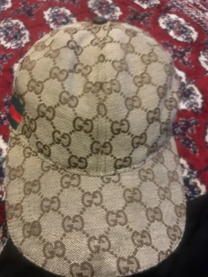 Gucci keps med GG-mönster - Snygg keps från Gucci med klassiskt GG-monogram i beige och brunt. Kepsen har böjd skärm och en grön-röd rand på sidan. Tillverkad i canvasmaterial med exklusiv känsla. Perfekt för dig som vill sticka ut med en ikonisk accessoar.