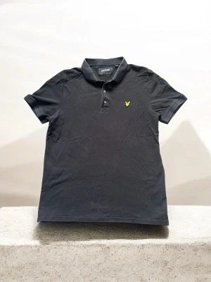 Svart piké från Lyle & Scott - Klassisk svart pikétröja från Lyle & Scott med korta ärmar och gul logga på bröstet. Tröjan har krage och knappar framtill. Tillverkad i mjuk bomull för en bekväm känsla och stilren look.