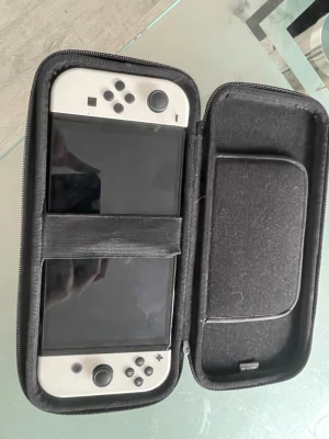 Nintendo Switch OLED vit med fodral - Nintendo Switch OLED i vit färg med matchande vita Joy-Con-kontroller. Konsolen kommer i ett robust svart skyddsfodral med Nintendo Switch-logga och prägling. Perfekt för gaming på språng och enkel att ta med sig tack vare det praktiska fodralet.