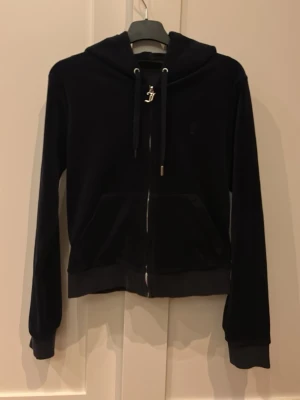 Mörkblå velour hoodie från Juicy Couture - Mörkbå hoodie i mjuk velour från Juicy Couture. Dragkedja framtill, två fickor och huva med snören som har metalländar och ett J-hänge. Perfekt för en chill och trendig look. Den saknar en metall bit på snöret.💕