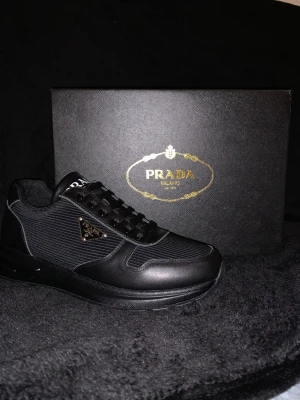 Svarta Prada sneakers i läder och textil - Snygga svarta sneakers från Prada med stilren design i läder och textil. Skon har svarta skosnören, diskret logga på sidan och en chunky sula. Perfekt för dig som gillar exklusiva och trendiga skor.