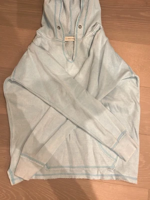 Ljusblå hoodie från Ocean Drive - Ljusblå hoodie från Ocean Drive med trycket 'Nomad Surf Shop' och en hibiskusblomma på bröstet. Hoodien har huva med knappar, magficka och synliga sömmar i blått. Tillverkad i mjukt bomullsmaterial, perfekt för chill dagar.