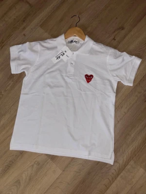 Vit pikétröja Comme des Garçons Play - Vit pikétröja från Comme des Garçons Play med klassisk krage, korta ärmar och knappar framtill. På bröstet sitter det ikoniska röda hjärta-märket. Tillverkad i mjuk bomull för en skön och clean look.