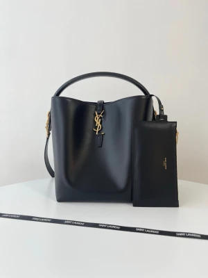 Svart bucket bag från Saint Laurent - Snygg svart bucket bag från Saint Laurent i slätt läder med guldfärgade detaljer och ikoniskt YSL-emblem framtill. Väskan har ett topphandtag, justerbar axelrem och medföljande svart clutch. Perfekt för dig som vill ha en stilren och lyxig handväska.