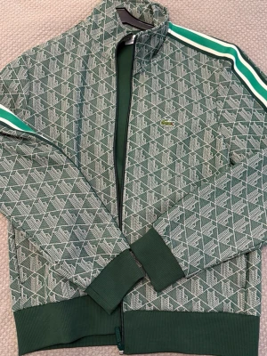 Grön mönstrad track jacket Lacoste - Snygg grön track jacket från Lacoste med vitt och grönt mönster över hela tröjan. Helt oanvänd med prislapp kvar, köpt från Lacoste butiken. Nypris 950. 