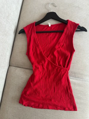 Ribbad röd omlottopp - Snygg ribbad röd topp med omlottdesign och v-ringning. Ärmlös modell som sitter figurnära och har en stretchig känsla. Storlek S