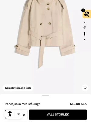 Beige kort trenchcoat med ståkrage - Trendig beige trenchcoat med ståkrage, dubbelknäppning och bälte i midjan. Jackan har snygga knappar och justerbara ärmslut på ärmen. säljer för att jag vill ha en grön och jag köpte denna i butik så kan inte lämna tillbaka, jackan har kvar prislappen och aldrig använd 