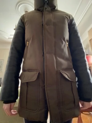 Brun parkas från Mackage XS - Snygg brun parkas från Mackage med dragkedja och knappar framtill, två stora fickor med lock och två snedställda fickor med guldfärgade dragkedjor. Jackan har hög krage och långa ärmar, perfekt för kalla dagar.