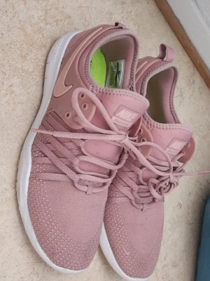 Rosa Nike sneakers med meshdetaljer - Säljer ett par snygga rosa Nike sneakers med stickad mesh på ovandelen och vita sulor. Skorna har klassisk Nike-logga på plösen och sidan samt gröna innersulor med svart text. Perfekta för dig som gillar sportig och trendig stil.