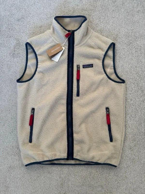 Beige fleeceväst från Patagonia - Snygg beige fleeceväst från Patagonia med marinblå detaljer och röda dragkedjor. Västen har hög krage, tre fickor med dragkedja och är tillverkad i mjukt fleece-material. Perfekt för lager-på-lager och outdoor-vibe.