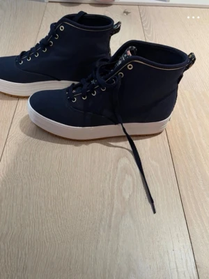 Mörkblå höga sneakers från Keds - Snygga mörkblå sneakers från Keds i samarbete med Rifle Paper Co. Skorna har högt skaft, vita sulor och guldfärgade öljetter. Insidan har ett blommigt mönster och textilmaterial. Perfekta för dig som vill ha en unik och stilren look.