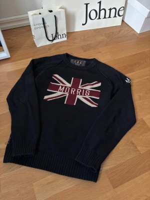 Mörkblå/svart stickad tröja Morris - Snygg mörkblå/svart stickad tröja från Morris med Union Jack-flagga i vinrött, vitt och grått på bröstet samt Morris-logga. Tröjan har rund halsringning, ribbade muddar och är perfekt för dig som gillar klassisk brittisk stil.