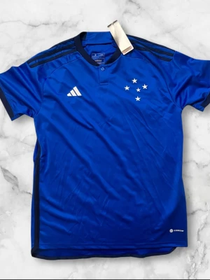 Blå Adidas fotbollströja Cruzeiro - Snygg blå fotbollströja från Adidas med Cruzeiro-stjärnor på bröstet och klassiska Adidas-ränder på axlarna. Tillverkad i lätt och ventilerande funktionsmaterial, perfekt för träning eller match. Kortärmad modell med knapp i halsen och diskreta svarta detaljer.
