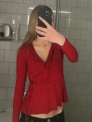 Röd omlottblus med volang - Snygg röd omlottblus med volangkant längs ringningen och knytband i sidan. Blusen har v-ringning och lång ärm, samt lätt utsvängd nederdel som ger en feminin siluett. Perfekt att styla med jeans eller kjol. Säljer den då den är för stor för mig och inte kommer till användning, kontakta mig om ni har några frågor💕