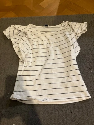 Vit randig topp med volangärmar - Snygg vit topp med tunna svarta ränder och volangiga korta ärmar. Toppen har en avslappnad passform och rund halsringning. Perfekt för dig som gillar en enkel men trendig stil.