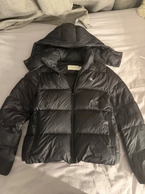 Svart pufferjacka från Calvin Klein - Snygg svart pufferjacka från Calvin Klein med avtagbar huva och dragkedja framtill. Säljs för 750kr då den är köpt för 1500kr. Jackan är i storlek M men passar även mig som har S! Jackan har två stora fickor med dragkedja och diskret CK-logga på bröstet. Perfekt för kalla vinterdagar och har en modern, boxig passform. Passar även passformen män. 