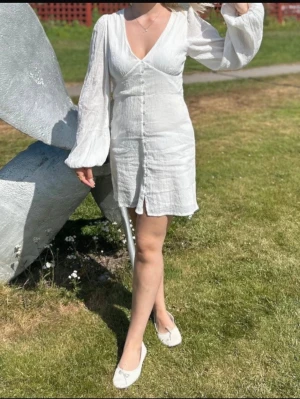 Vit kort klänning  - Säljer en vit kort klänning från Nelly med v-ringning, långa puffärmar och knappar framtill. Använd 1 gång 