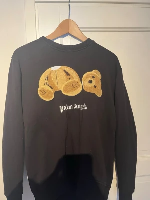 Svart sweatshirt från Palm Angels - Svart sweatshirt från Palm Angels med ikoniskt nallebroderi på bröstet och vit logotyptext. Tröjan har rund halsringning och ribbade muddar. Perfekt för dig som gillar streetwear och vill sticka ut med en unik detalj. 