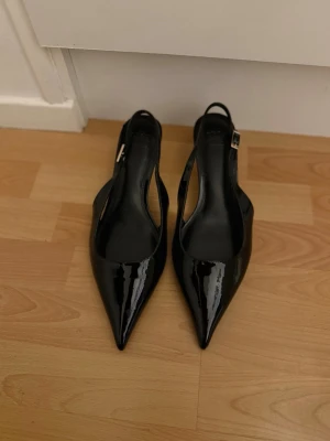 Svarta lackpumps med öppen häl - Säljer ett par svarta pumps i glansigt lackmaterial med spetsig tå och öppen häl. Skorna har en smal rem med spänne runt hälen och klassisk siluett. Perfekta för dig som gillar stilrena och trendiga skor. 