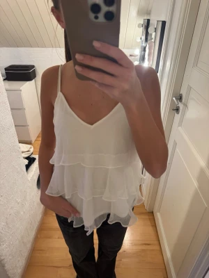 Vit volangig linnetopp - Säljer en söt vit linnetopp med tunna axelband och flera lager volanger. Toppen har v-ringning och ett luftigt, flowy material som passar perfekt till sommaren.