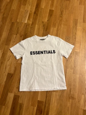 Vit Essentials T-shirt Fear of God - Säljer en clean vit T-shirt från Fear of God Essentials med stor svart logga på bröstet. Klassisk rund hals och korta ärmar. Tillverkad i mjuk bomull för skön känsla och enkel stil. Perfekt för dig som gillar streetwear och minimalistisk design.