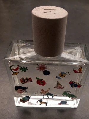 Parfymflaska med lekfulla motiv från populära Maison Matines doft Pom Pom! - Unik parfymflaska i genomskinligt glas med färgglada, lekfulla illustrationer som pryder framsidan. Flaskan har en fyrkantig form och ett stort, ljust korklock i naturmaterial. Perfekt för dig som gillar kreativa och personliga detaljer på ditt sminkbord. Helt ny med ordinarie pris 660 kr.