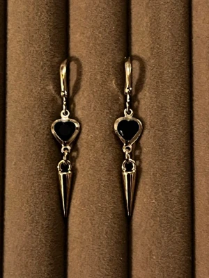 Silverörhängen med svarta hjärtan - Snygga örhängen i silverfärg med svarta hjärtformade detaljer och spetsiga hängen. Örhängena har en modern och edgy design med klicklås och metallisk finish. Perfekta för dig som gillar statement-smycken med en unik twist.