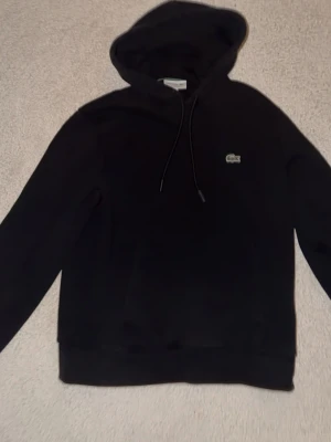 Svart hoodie från Lacoste - Snygg svart hoodie från Lacoste med klassisk krokodillogga på bröstet. Hoodien har dragsko i huvan och är gjord i mjukt bomullsmaterial. Perfekt för en chill och stilren look.