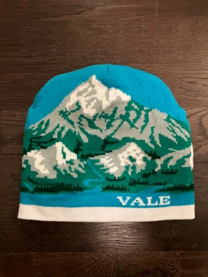 Turkos mössa med bergsmotiv från Vale - Säljer en turkos mössa från Vale med ett snyggt bergsmotiv i vitt, grönt och grått, perfekt för att vistas ute i naturen eller bara kunna vara ute och njuta utan att frysa. Mössan har en vit kant längst ner och är stickad i mjukt material, perfekt för kalla dagar. Trycket är detaljerat och ger en cool outdoor-vibe. 