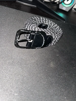 Svart och gråt flätat bälte - Snyggt svart och gråt flätat bälte med metallspänne. Passar perfekt till jeans eller byxor. Flätad design gör det enkelt att justera längden. Perfekt accessoar för att lyfta din outfit.