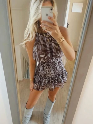 Brun mönstrad kortklänning med volang - Säljer en snygg kortklänning med brunt och vitt zebramönster. Klänningen har ärmlös design, smockad midja och volang nertill. Perfekt för dig som gillar trendiga och unika plagg. Materialet känns lätt och luftigt.