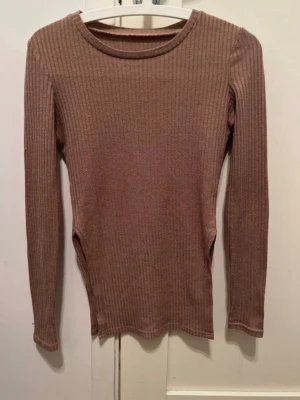 Brun ribbad långärmad tröja - Säljer en brun ribbad långärmad tröja med rund halsringning och slit vid sidan av magen. Tröjan är figurnära och har en enkel, stilren design som passar perfekt till jeans. Materialet är mjukt och stretchigt för en bekväm känsla hela dagen.