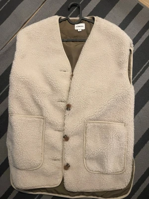 Beige teddyväst från Kappahl - Mysig beige teddyväst från Kappahl med två stora framfickor och knäppning med träknappar. Västen har en klassisk rak passform och är fodrad på insidan. Perfekt för lager på lager och ger en trendig look.