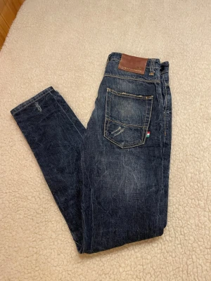 Adrian Hammod Jeans  - Snygga mörkblå slim fit jeans från Adrian Hammond, Jeans med slitningar och detaljer på både fram- och baksida. Klassisk femficksmodell med läderpatch bak i midjan och liten italiensk flaggdetalj vid bakfickan. Perfekta för en trendig och avslappnad look.