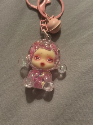 Skullpanda nyckelring med vätska i och glitter - Nyckelring med genomskinlig figur fylld med rosa och vita blommor samt glitter. Figuren har små armar och ben, och nyckelringen har även en rosa klocka. Perfekt som accessoar till väskan eller nycklarna.