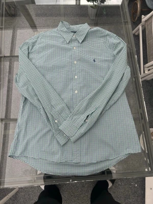 👕 Ralph Lauren skjorta – Custom Fit – XL - Rutig skjorta i grönt, blått och vitt från Ralph Lauren. Klassisk och stilren modell i Custom Fit – perfekt för både vardag och finare tillfällen. Passar snyggt till jeans, chinos eller kavaj.  ✨ Info: 	•	Märke: Ralph Lauren 	•	Passform: Custom Fit 	•	Storlek: XL 	•	Färg: Grön / Blå / Vit 	•	Mönster: Rutig (gingham) 	•	Material: Bomull 	•	Skick: Använd  ⚠️ Finns mindre slitage/hål på ena ärmen (se bild). Syns mest vid närmare granskning.  Tidlös preppy-stil och ett riktigt bra plagg till garderob