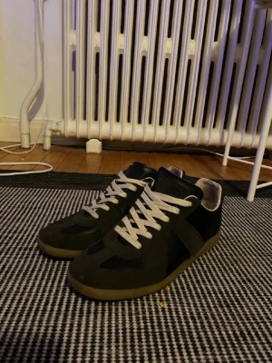 Maison Margiela gats  - Stilrena och tidlösa sneakers i svart mocka/skinn med beige sula och vita snören. Den ikoniska siluetten gör dem enkla att matcha – funkar lika bra till jeans, kostymbyxor eller mer dressade fits. Diskret men statement för den som gillar clean designer pieces.  Skick: Väldigt bra skick, sparsamt använda (se bilder). Färg: Brun med gum sole Nypris: ca 7-8000 kr Säljes pga används för lite.  Skicka DM vid frågor eller fler bilder ✨