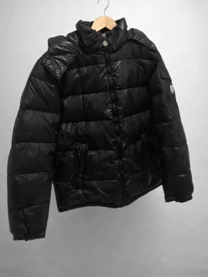 Svart pufferjacka  - Snygg svart pufferjacka från  med hög krage och dragkedja framtill. Jackan har två fickor med dragkedja och  på . Tillverkad i glansigt material och har en klassisk quiltad design som håller värmen under kalla dagar.