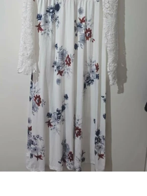 Vit blommig långklänning med spets - Vit långklänning med blommigt mönster i blått och rött. Klänningen har långa spetsärmar med vackra detaljer och en luftig passform. Perfekt för sommarens alla tillfällen och ger en romantisk känsla.