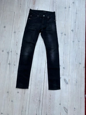 Svarta jeans ifrån Tiger of Sweden - Säljer ett par svarta jeans från Tiger of Sweden med klassisk femficksdesign och diskreta slitningar. Jeansen har raka ben och knappgylf. Perfekta för dig som gillar en clean och stilren look till vardags. Materialet är troligtvis bomull med lite stretch.