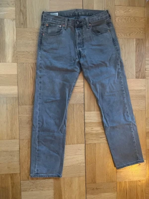 Grå raka jeans från Levi's - Säljer ett par grå jeans från Levi's med klassisk femficksmodell och raka ben. Jeansen har normal midja, bälteshällor och knappgylf. Materialet är slitstark denim i bomull. Perfekta för en avslappnad och stilren look.