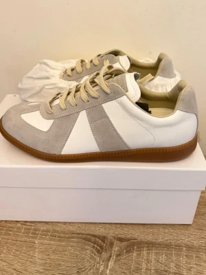 Maison Margiela Replica sneakers vit/grå - Maison Margiela Replica sneakers i vitt och ljusgrått med beige snörning och gummisula. Ovandelen är i en mix av mocka och läder, klassisk låg modell med retrovibbar. Perfekta för dig som gillar stilrena och tidlösa sneakers.