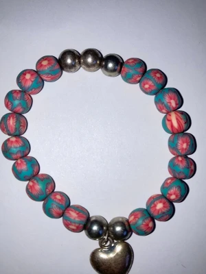 Blommigt pärlarmband med hjärthänge - Färgglatt armband med runda pärlor i turkos och rosa med blommönster, kombinerat med silvriga pärlor. Armbandet har ett silverfärgat hjärthänge som detalj. Perfekt accessoar för dig som gillar unika och lekfulla smycken.